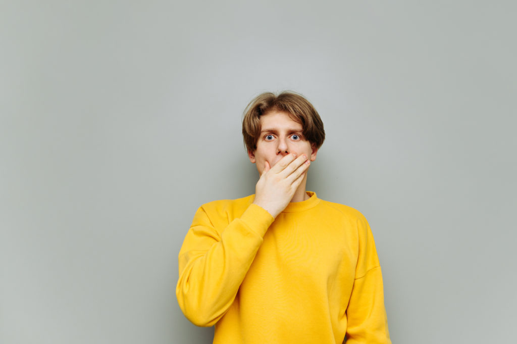 Shocked young man in casual clothes covers his face with his hand and looks at the camera with a surprised face isolated on a gray background.